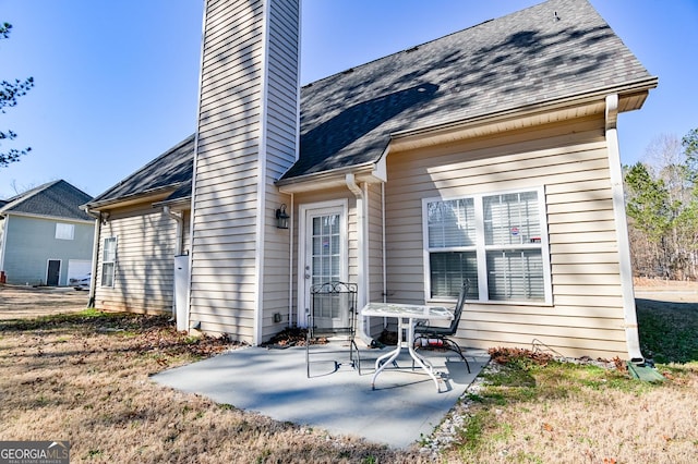 back of house with a shingled roof, a patio, and a chimney