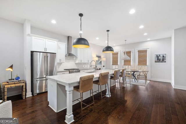 kitchen with high quality appliances, white cabinets, wall chimney range hood, an island with sink, and decorative light fixtures