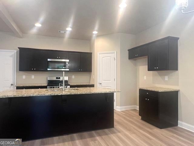 kitchen featuring a kitchen island with sink, dark cabinets, light wood-style floors, appliances with stainless steel finishes, and light stone countertops