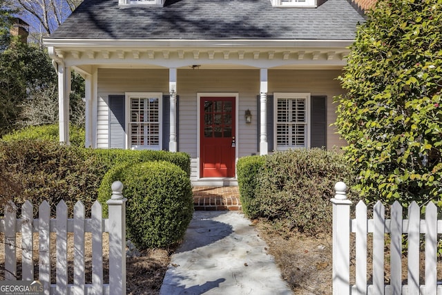 view of exterior entry with covered porch, roof with shingles, and fence