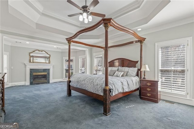 carpeted bedroom with a tray ceiling, a fireplace, crown molding, a ceiling fan, and baseboards