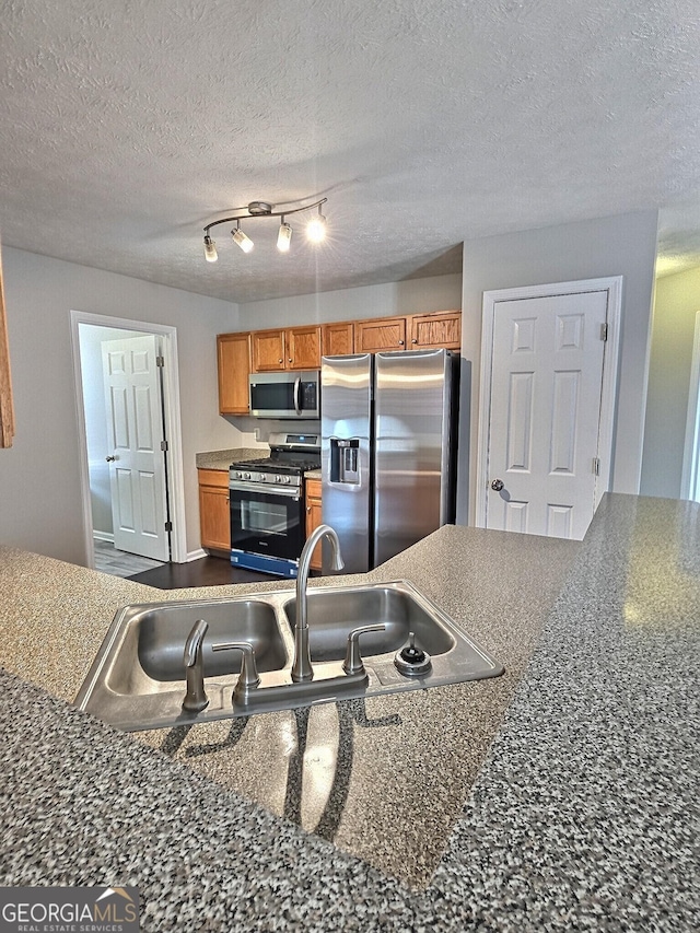 kitchen with brown cabinetry, dark countertops, stainless steel appliances, a textured ceiling, and a sink