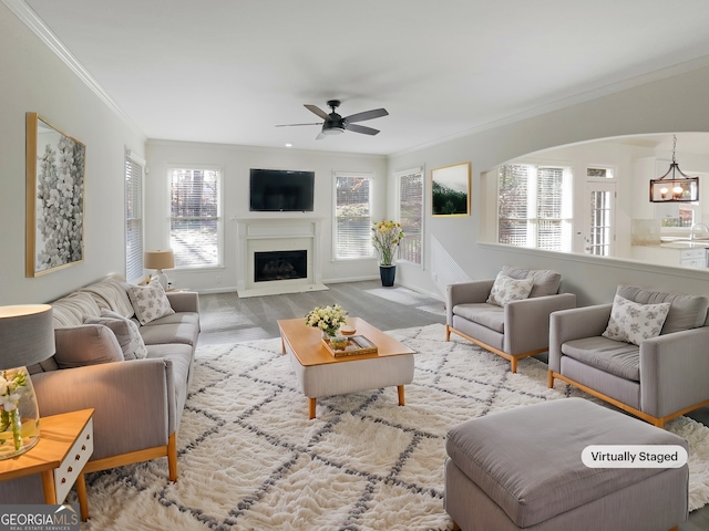 living area featuring arched walkways, ceiling fan with notable chandelier, a fireplace, wood finished floors, and crown molding