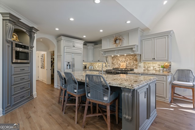 kitchen with arched walkways, a kitchen island with sink, light stone counters, and light wood-style flooring