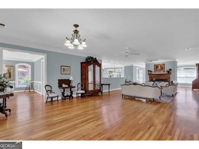 living room featuring baseboards, ornamental molding, light wood-type flooring, a fireplace, and ceiling fan with notable chandelier