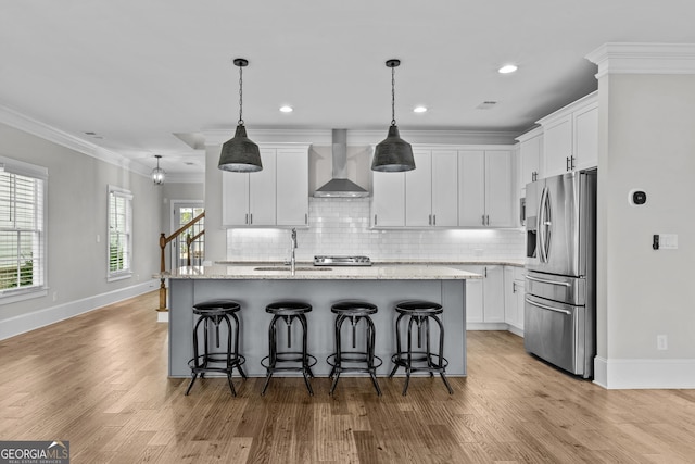 kitchen with crown molding, stainless steel fridge with ice dispenser, a kitchen island with sink, white cabinets, and wall chimney exhaust hood