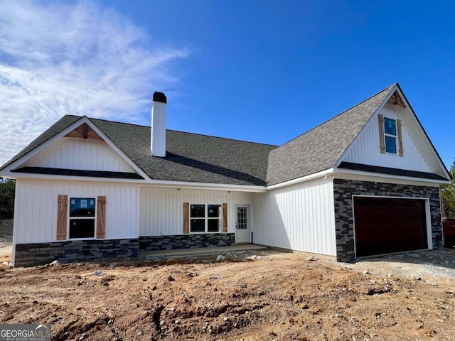 modern farmhouse featuring stone siding, driveway, a chimney, and roof with shingles