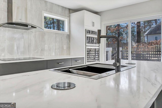 kitchen with light stone countertops, black electric cooktop, wall chimney range hood, white cabinetry, and a sink