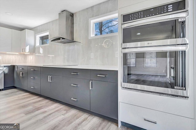 kitchen featuring double oven, white cabinets, light countertops, decorative backsplash, and wall chimney exhaust hood