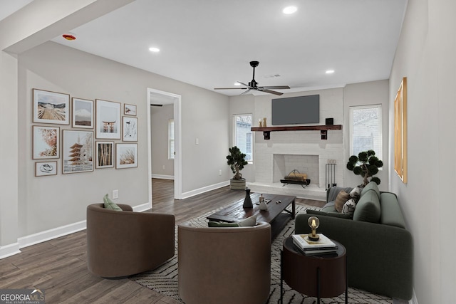 living area featuring dark wood-type flooring, a brick fireplace, baseboards, and a ceiling fan