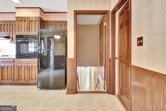 kitchen with a wainscoted wall, brown cabinetry, extractor fan, black appliances, and wallpapered walls