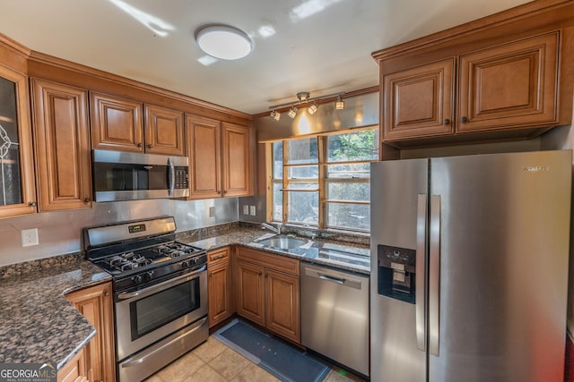 kitchen with stainless steel appliances, a sink, brown cabinets, dark stone countertops, and glass insert cabinets