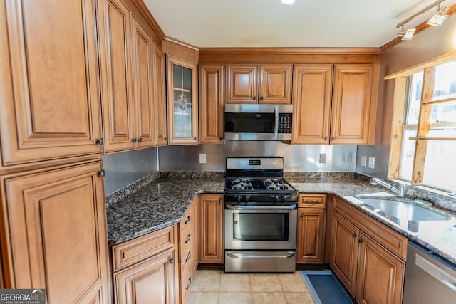 kitchen featuring appliances with stainless steel finishes, brown cabinetry, glass insert cabinets, a sink, and dark stone countertops