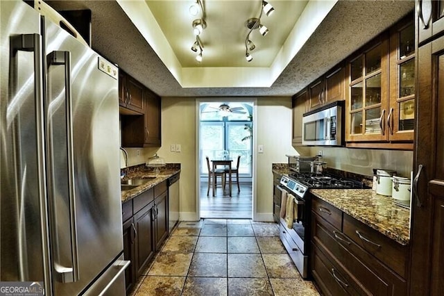 kitchen with a raised ceiling, ceiling fan, appliances with stainless steel finishes, dark stone countertops, and a sink