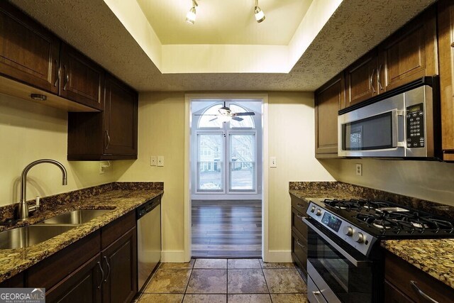 kitchen with a ceiling fan, appliances with stainless steel finishes, dark stone countertops, a tray ceiling, and a sink