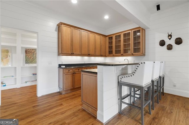 kitchen featuring a peninsula, wood finished floors, brown cabinetry, dark countertops, and glass insert cabinets