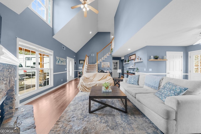 living area featuring a towering ceiling, ceiling fan, a stone fireplace, wood finished floors, and stairs