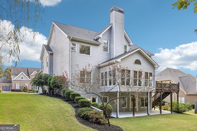 rear view of house with roof with shingles, a chimney, a lawn, a patio area, and a deck