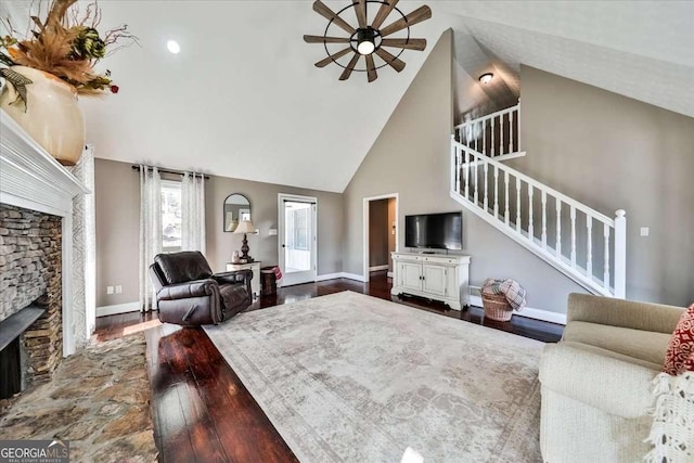 living room with high vaulted ceiling, a stone fireplace, wood finished floors, baseboards, and stairs