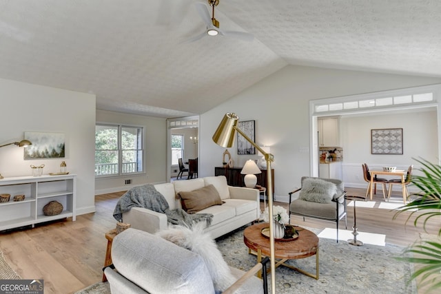 living area featuring a textured ceiling, light wood-style flooring, a ceiling fan, baseboards, and vaulted ceiling