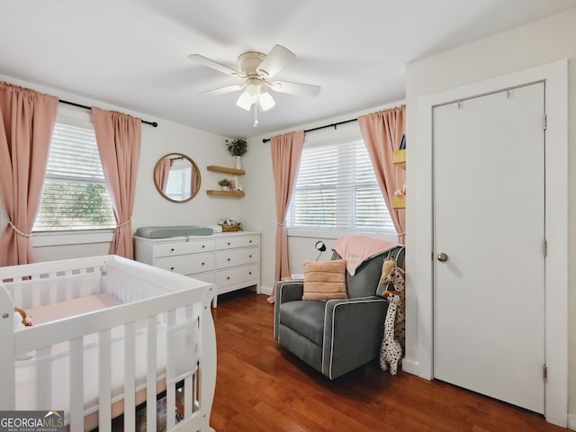 bedroom with a nursery area, multiple windows, dark wood finished floors, and a ceiling fan