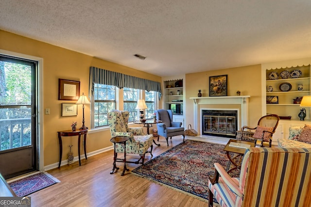 living area with built in features, light wood-style floors, a glass covered fireplace, a textured ceiling, and baseboards