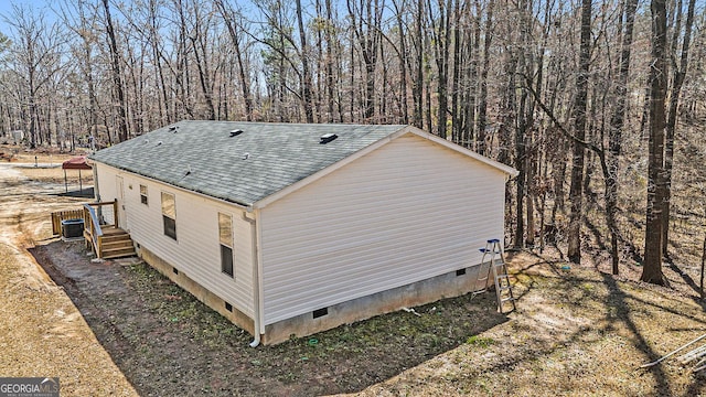 view of side of home with central AC, crawl space, a forest view, and roof with shingles