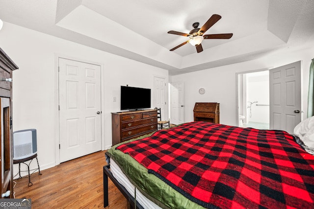 bedroom with baseboards, a raised ceiling, a ceiling fan, ensuite bathroom, and light wood-style floors