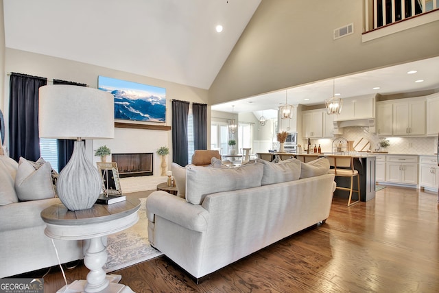 living area with a fireplace with raised hearth, high vaulted ceiling, a chandelier, wood finished floors, and visible vents