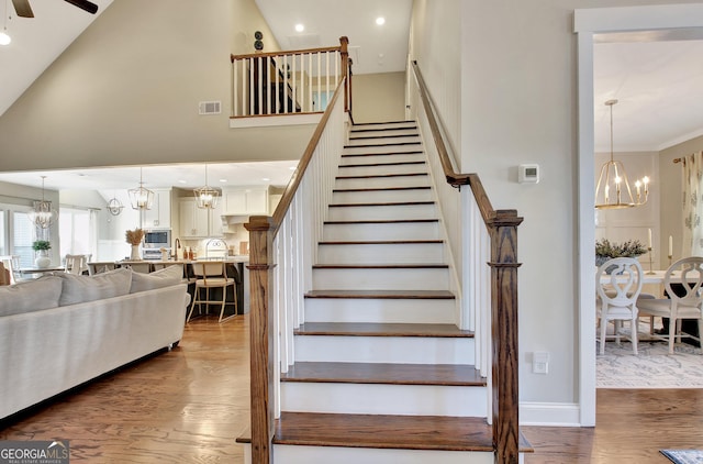 staircase featuring high vaulted ceiling, ceiling fan with notable chandelier, visible vents, and wood finished floors