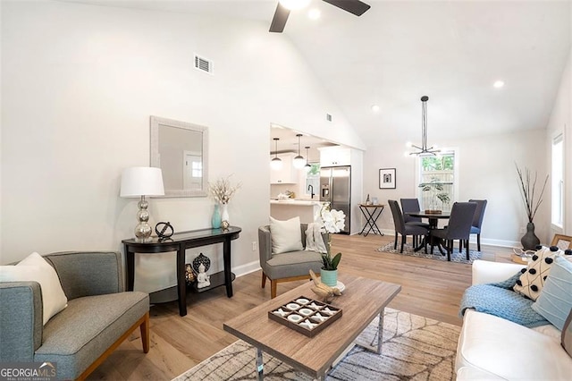 living room with high vaulted ceiling, light wood-style flooring, ceiling fan with notable chandelier, visible vents, and baseboards