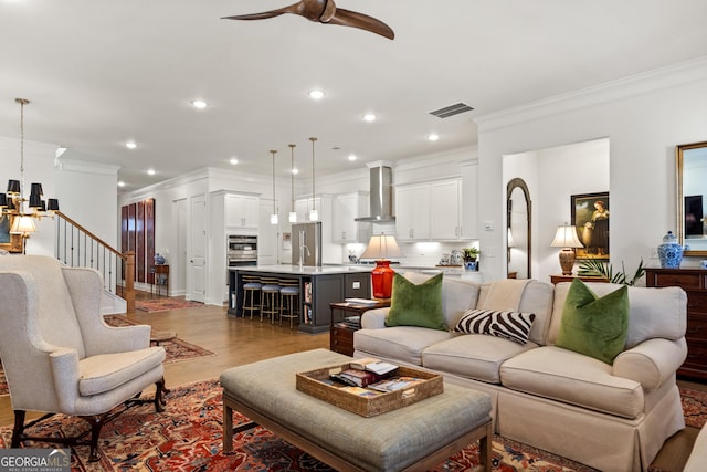 living room featuring recessed lighting, visible vents, light wood-style flooring, ornamental molding, and ceiling fan with notable chandelier