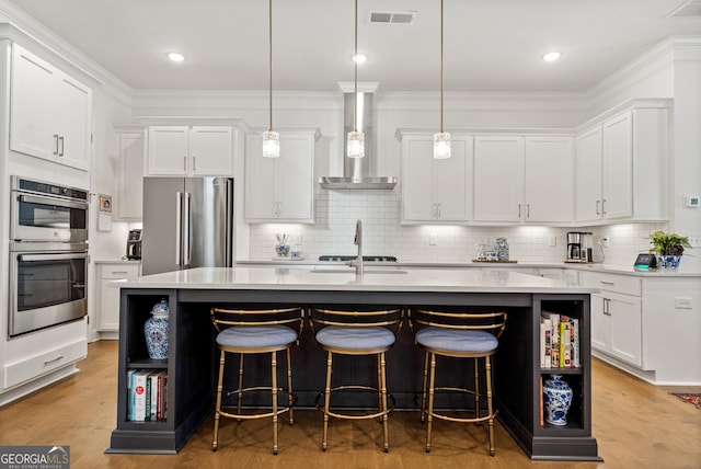 kitchen with visible vents, wall chimney exhaust hood, appliances with stainless steel finishes, a kitchen island with sink, and light countertops