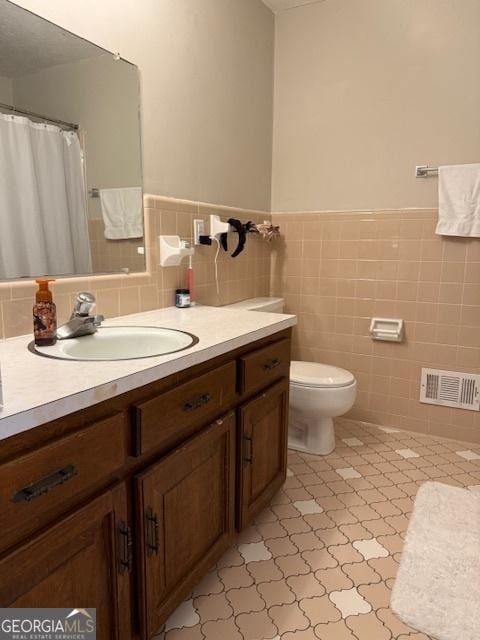 bathroom with toilet, a wainscoted wall, vanity, visible vents, and tile walls