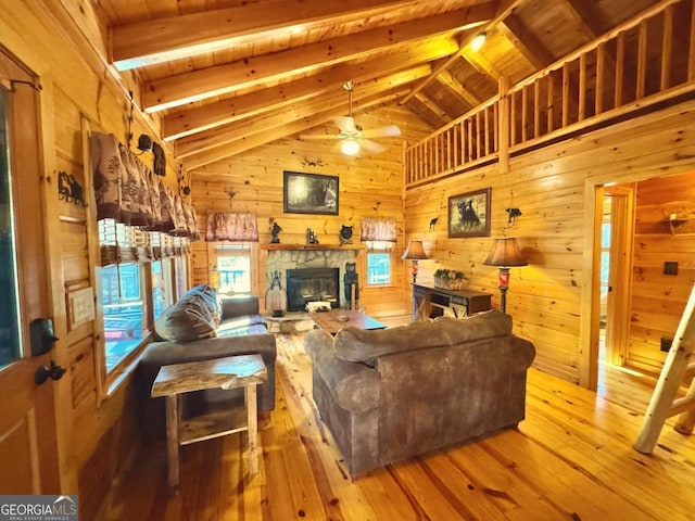 living room with beam ceiling, light wood finished floors, wood walls, a stone fireplace, and ceiling fan