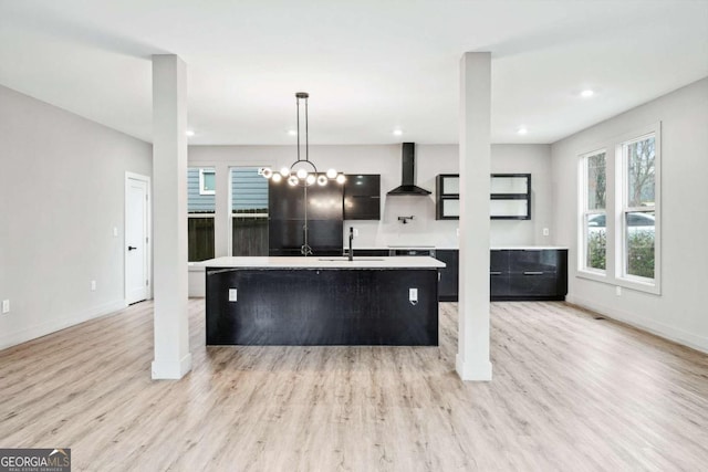 kitchen featuring a sink, light countertops, hanging light fixtures, wall chimney range hood, and a center island with sink