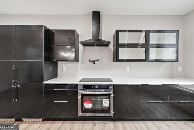 kitchen featuring wall chimney exhaust hood, light countertops, stainless steel oven, and black electric cooktop