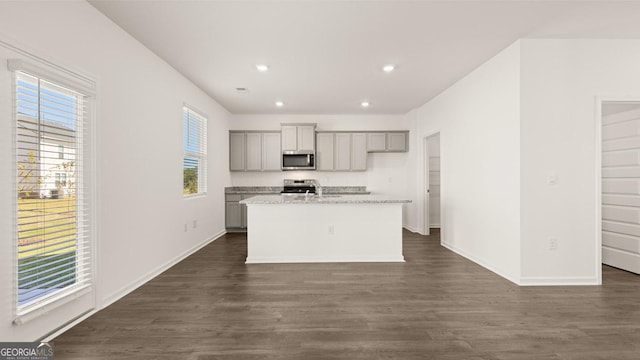 kitchen with light stone counters, a kitchen island with sink, stainless steel appliances, gray cabinets, and dark wood finished floors