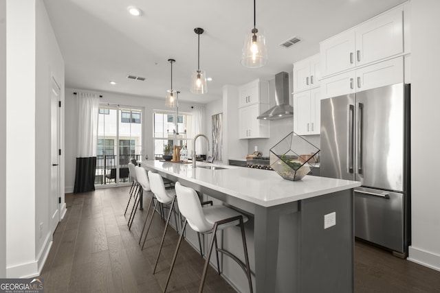 kitchen featuring light countertops, white cabinets, high quality fridge, an island with sink, and wall chimney exhaust hood