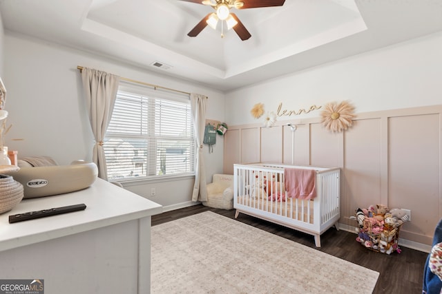 bedroom with a crib, a decorative wall, visible vents, a tray ceiling, and dark wood finished floors