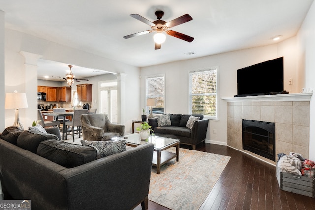 living area featuring decorative columns, baseboards, dark wood finished floors, a ceiling fan, and a tiled fireplace