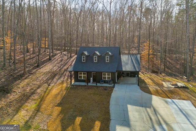 cape cod house featuring covered porch, a front lawn, a wooded view, and concrete driveway