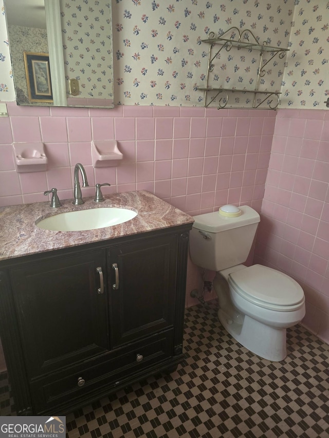 half bathroom with toilet, a wainscoted wall, tile walls, vanity, and wallpapered walls