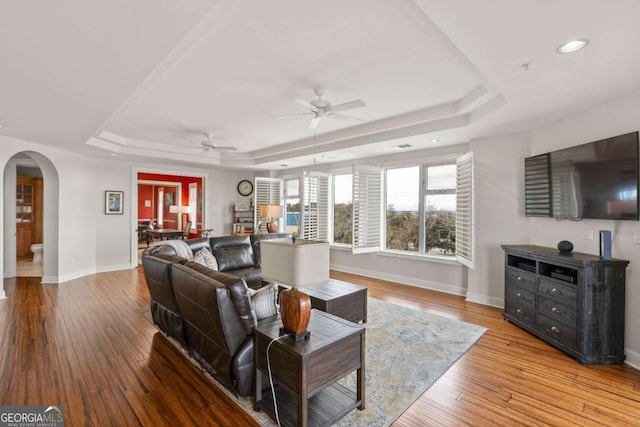 living room with a ceiling fan, a tray ceiling, arched walkways, wood-type flooring, and baseboards