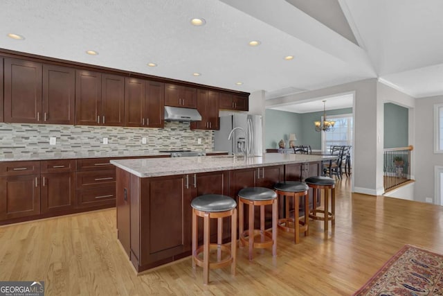 kitchen featuring a center island with sink, stainless steel refrigerator with ice dispenser, backsplash, light stone countertops, and under cabinet range hood