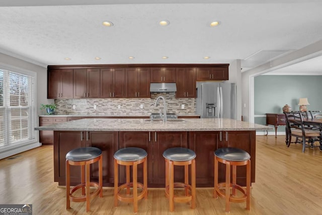 kitchen featuring light stone counters, under cabinet range hood, stainless steel refrigerator with ice dispenser, and an island with sink