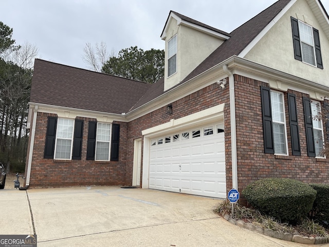 view of side of home featuring concrete driveway, brick siding, roof with shingles, and stucco siding