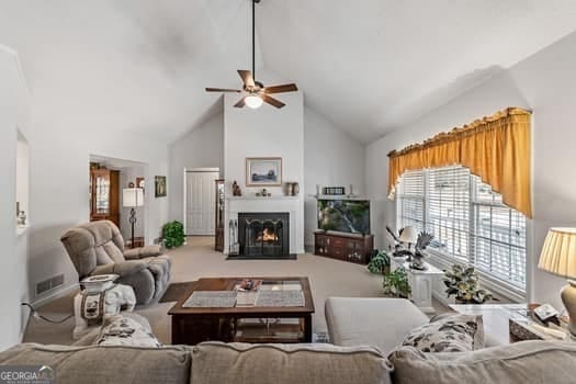 living area with a warm lit fireplace, high vaulted ceiling, ceiling fan, visible vents, and carpet