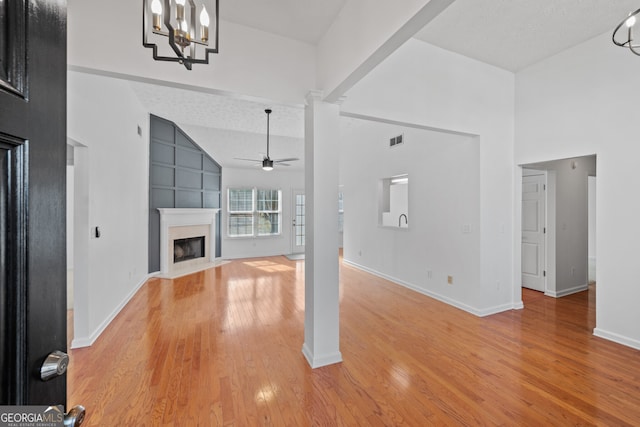 unfurnished living room with a fireplace, visible vents, light wood-type flooring, ornate columns, and ceiling fan with notable chandelier