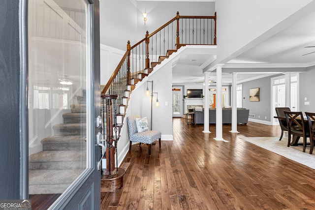 stairway with a high ceiling, a fireplace, ornate columns, wood-type flooring, and crown molding